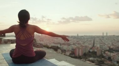 Young woman in bodysuit practices yoga at sunrise at viewpoint. Girl raises her hands up while sitting in lotus position. Back view