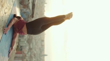 VERTICAL VIDEO: Woman doing yoga outdoors lying on mat with legs up in sportswear