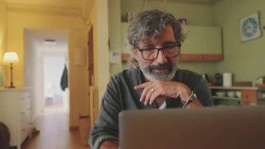 Elderly positive man typing working on laptop sitting in home kitchen
