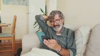 Evde koltukta otururken cep telefonu kullanan yaşlı pozitif bir adam.