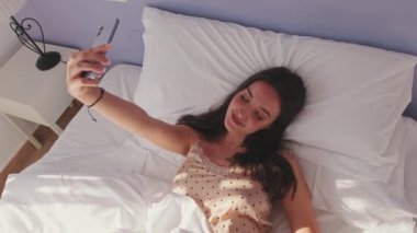 Young woman using her smart phone and lying in bed in the morning. Girl takes morning selfie on smartphone. Top view