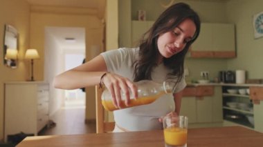 Young woman pouring orange juice into glass while sitting in the kitchen