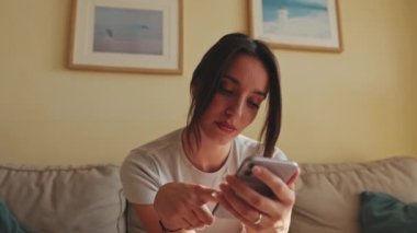 Close up, young woman using mobile phone for texting and scrolling while sitting on sofa at home
