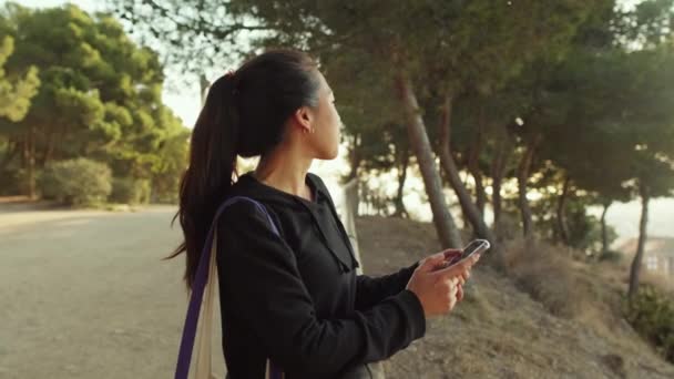 若い女性は彼女の手の中に携帯電話で公園内のパスに立って 周りを見回します — ストック動画