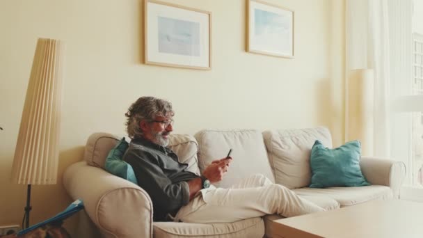高齢者はソファに座って携帯電話を使って家のリビングルームでリラックスして — ストック動画