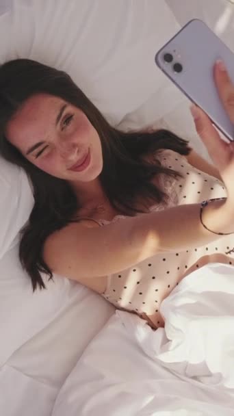 視覚的なビデオ 若い女性は彼女のスマートフォンを使用して 午前中にベッドに横たわっています 女の子はスマートフォンで朝の自撮りを取ります トップ表示 — ストック動画