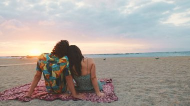 Kadın ve erkek sahilde birbirlerine sarılıp şafağı izliyorlar. Arka plan