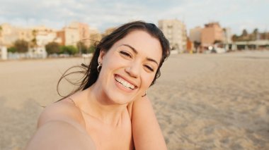 Genç bir kadın kumsalda oturuyor ve cep telefonu kullanıyor. Video çekiyor.