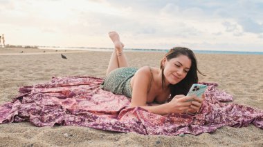 Kumsalda dinlenen ve cep telefonu kullanan gülümseyen genç bir kadın.