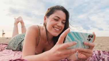 Genç bir kadın sahilde dinleniyor ve cep telefonu kullanıyor.