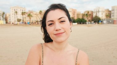 Genç kadın sahilde duruyor ve uzaklara bakıyor. Kız başını çevirir, kameraya bakar ve gülümser.