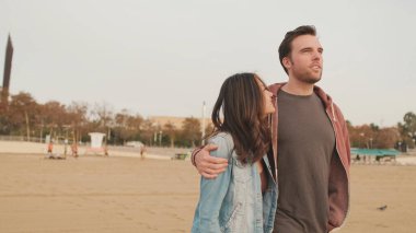 Birbirine aşık çiftler kumsalda yürürken birbirlerine sarılıyorlar.