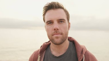 Sahilde dikilen ve kameraya bakan genç adamın yakın çekimi.