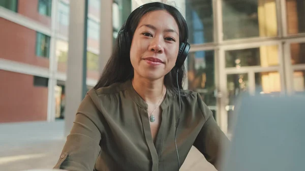 Young woman freelancer in headphones works on laptop, raises her head and smiles at camera