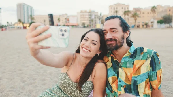 快乐的夫妻在沙滩上一边坐着一边用手机自拍 — 图库照片