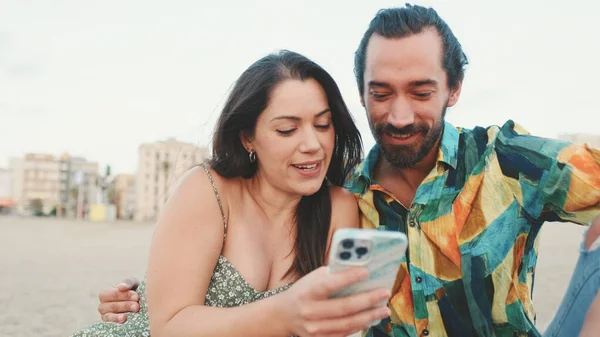 若いカップルが建物の背景にビーチに座っている間に携帯電話を使用して クローズアップ — ストック写真