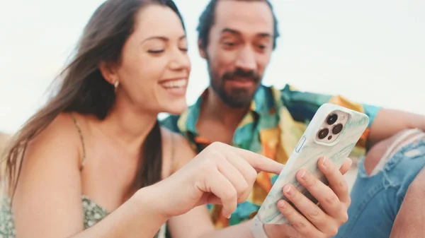 若いカップルが建物の背景にビーチに座っている間に携帯電話を使用して クローズアップ — ストック写真