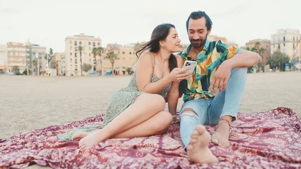 这对年轻夫妇坐在海滩上的时候用手机 背景是建筑物 特写镜头 — 图库照片
