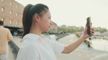 Cep telefonuyla deniz limanının fotoğrafını çeken genç bir kadın.
