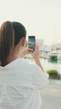 VERTICAL VIDEO: Young woman taking photo of the sea port on mobile phone. Back view