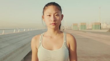 Asian girl in sports top walks forward along the promenade and looks at the camera