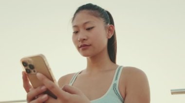 Close up, asian girl in sports top sits on the waterfront, uses smartphone. Backlight