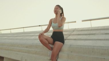 Asian girl in sports top sits on the embankment, talking on cellphone