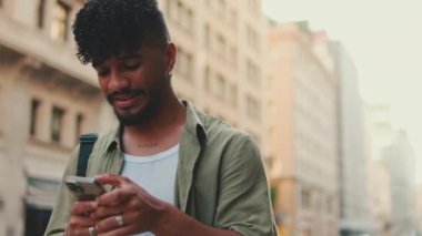 Zeytin rengi gömlek giymiş, sakallı, gülümseyen genç bir adam telefon kullanıyor.