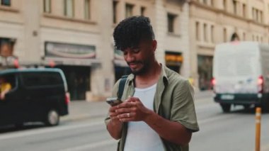 Young smiling man with beard dressed in an olive-colored shirt uses phone on an old city background. Guy reads the good news