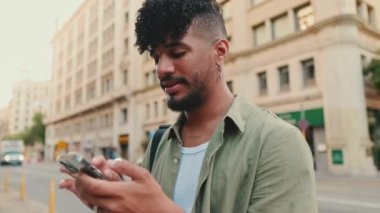 Close up, young happy man with beard dressed in an olive color shirt uses mobile phone while standing next to the road, raises his head and smiles at the camera