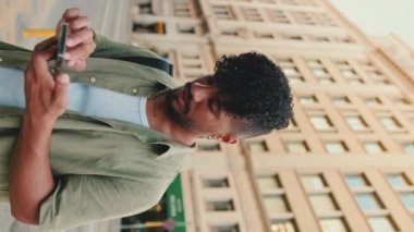 VERTICAL VIDEO: Young smiling man with beard dressed in an olive color shirt uses phone map app on the old city background