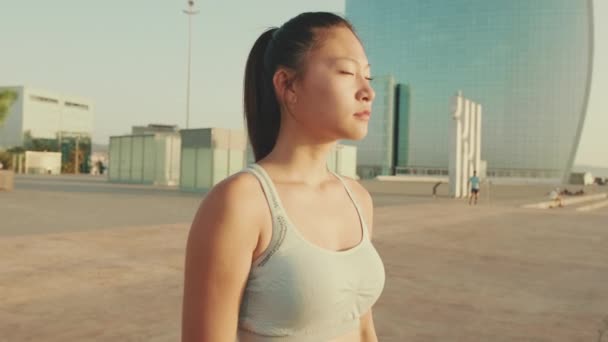 スポーツのトップのアジアの女の子は 現代的な建物の背景に立って 彼女の頭を回し カメラを見て — ストック動画