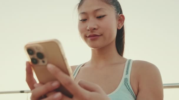 Close Smiling Asian Girl Sports Top Sits Waterfront Uses Smartphone — Stockvideo