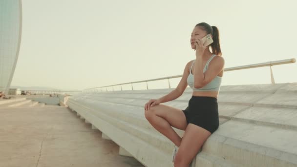 Asian Girl Sports Top Sits Embankment Talking Cellphone — Stok video