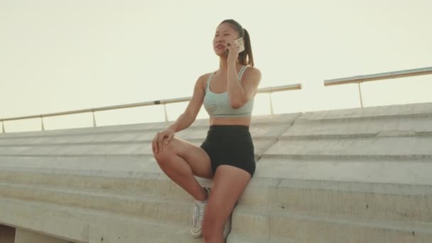 Asian Girl Sports Top Sits Embankment Talking Cellphone — Vídeo de Stock