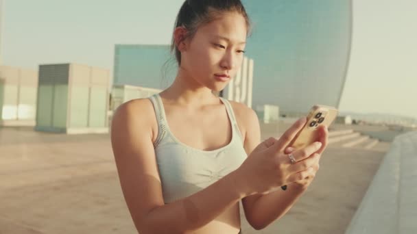 Close Smiling Asian Girl Sports Top Using Cellphone While Standing — ストック動画