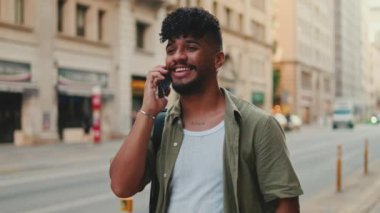 Young happy man with beard dressed in an olive-colored shirt is talking on cellphone while standing next to the road on the old city background