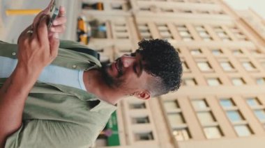 Yaklaş, zeytin rengi gömlek giymiş sakallı genç adam yolun kenarında dururken cep telefonu kullanıyor, başını kaldırıp kameraya gülümsüyor.