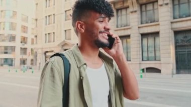 Young smiling man dressed in an olive color shirt walks down the street talking on the phone, the road and the old city background