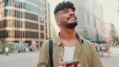 Young smiling man dressed in an olive-colored shirt stands with cellphone in his hand on modern building background