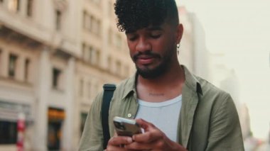 Close up, young smiling man with beard dressed in an olive-colored shirt uses phone on an old city background. Guy reads the good news