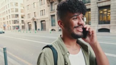Close up, young happy man with beard dressed in an olive-colored shirt is talking on cellphone walking down the street, the road and the old city background