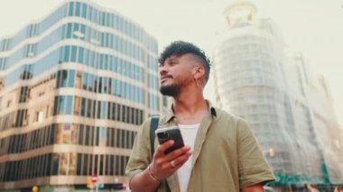 Young smiling man dressed in an olive-colored shirt stands with cellphone in his hand on modern building background