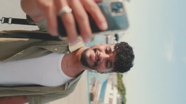 VERTICAL VIDEO: Young smiling man dressed in an olive-colored shirt takes selfie while standing on the waterfront in the port