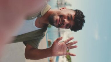 VERTICAL VIDEO: Close up, young smiling man dressed in an olive color shirt makes video call while standing on the waterfront in the port