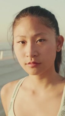 VERTICAL VIDEO: Close up, asian girl in sports top walks along the promenade and looks at the camera