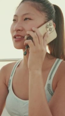VERTICAL VIDEO: Close up, asian girl in sports top sits on the embankment, talking on cellphone. Camera moves sideway to the left side. Backlight