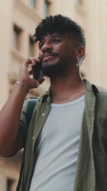 VERTICAL VIDEO: Young happy man with beard dressed in an olive-colored shirt is talking on cellphone while standing next to the road on the old city background