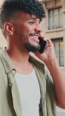 VERTICAL VIDEO: Young smiling man dressed in an olive color shirt walks down the street talking on the phone, the road and the old city background