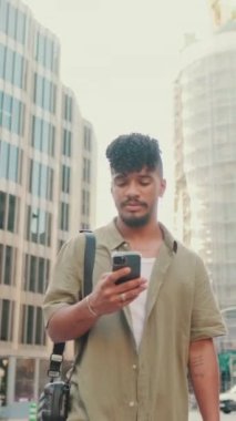 VERTICAL VIDEO: Young smiling man with beard dressed in an olive color shirt uses phone map app on the old city background